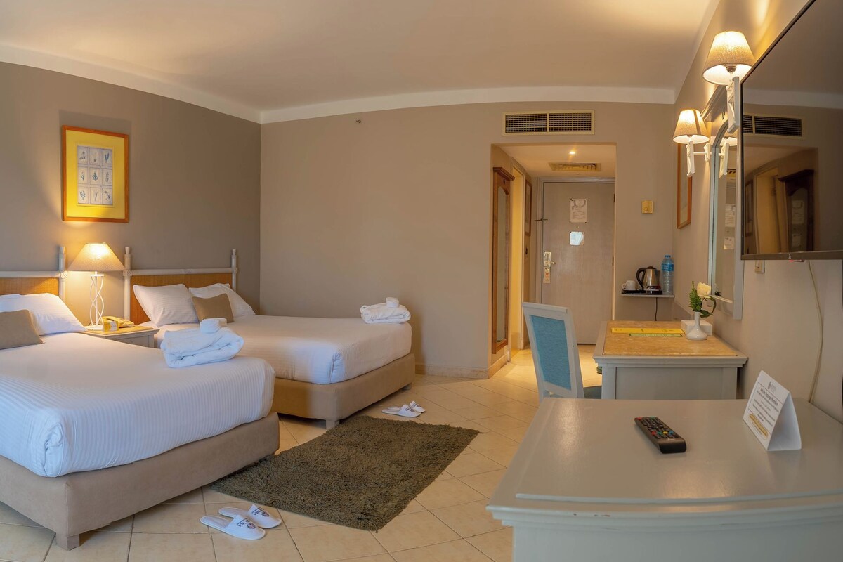 Une chambre d’hôtel avec deux lits, un coin salon comprenant une chaise et une table, une lampe de chevet et une petite table sur laquelle se trouvent un panier de fruits et des verres.