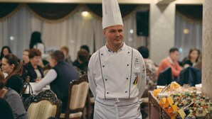 A chef in a white uniform with a tall hat standing in a room with a buffet table filled with various dishes. - Hotel Impero (Oradea)