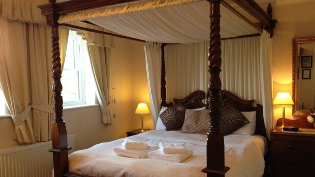 A four-poster bed with white linens and towels, a bedside table with a lamp, and a nightstand with a mirror and a remote control. - Ascot House (York)