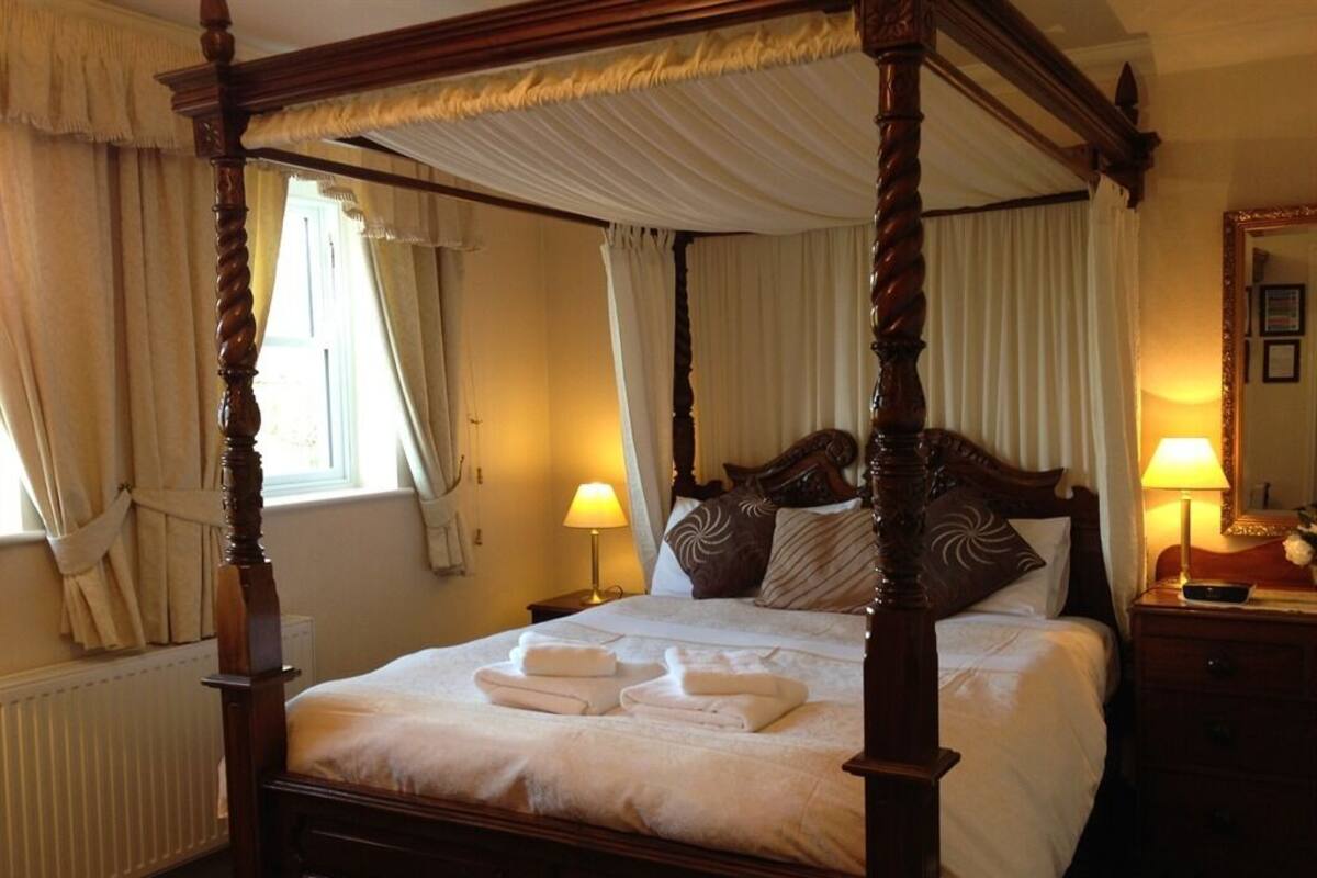 A four-poster bed with white linens and towels, a bedside table with a lamp, and a nightstand with a mirror and a remote control.