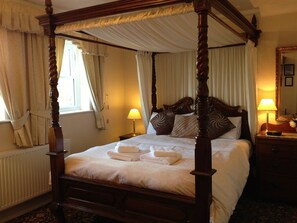 A four-poster bed with white linens and towels, a bedside table with a lamp, and a nightstand with a mirror and a remote control. - Ascot House (York)