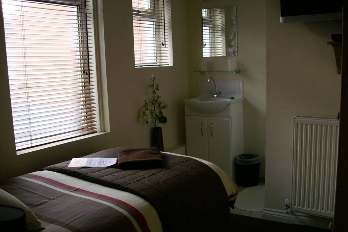 Un dormitorio con cama, mesita de noche, ventana con persianas, planta junto a la cama, baño con lavamanos y espejo, y un radiador.