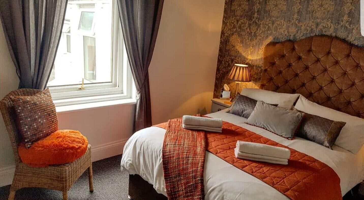 A hotel room with a large bed, a chair with an orange cushion, a nightstand with a lamp, and a window with curtains.