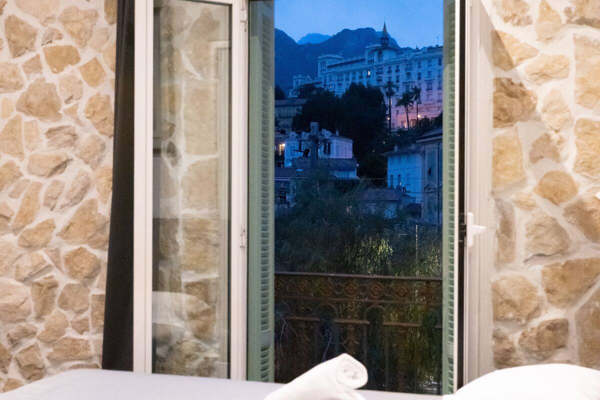 Habitación de hotel con pared de piedra, cama con ropa de cama blanca y vista de un paisaje urbano a través de un vidrio.