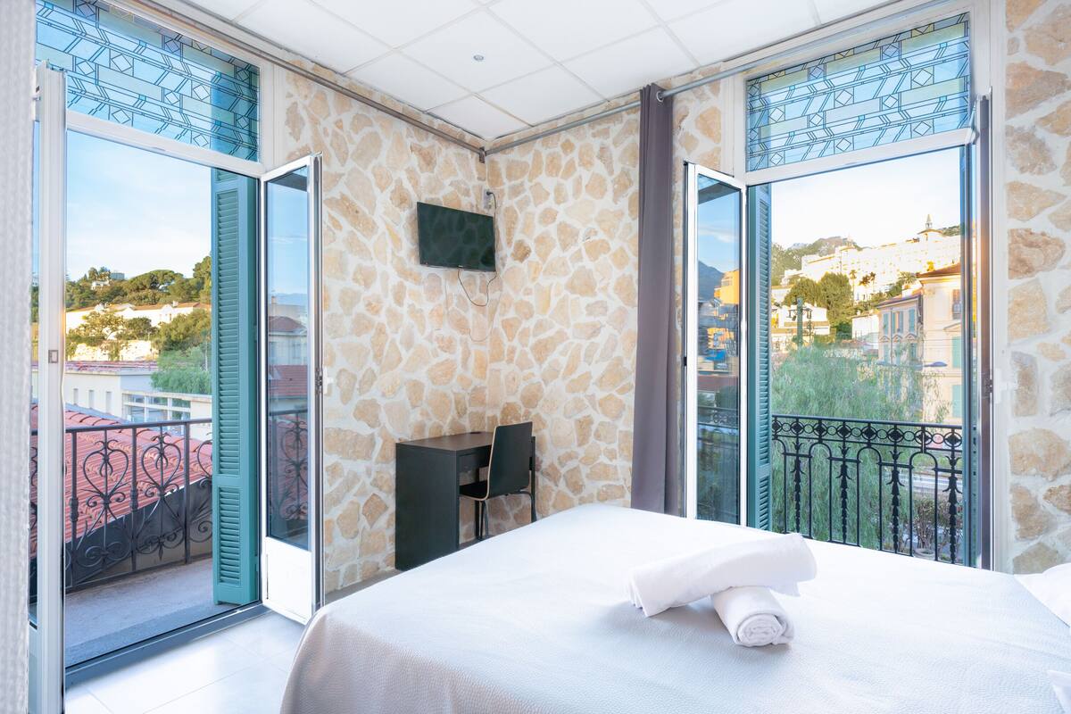 Habitación de hotel con cama, escritorio y silla. Tiene una televisión en la pared y un balcón con vista a la ciudad.