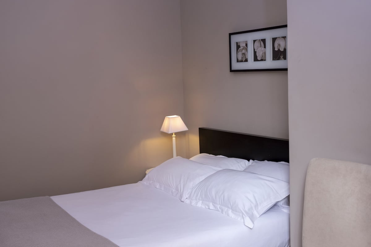 A neatly made bed with white linens and pillows, a bedside lamp, and a framed picture on the wall.