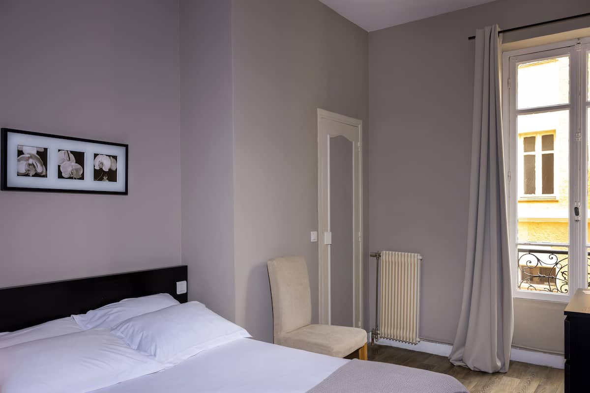 A hotel room with a bed, a chair, a radiator, and a window with curtains.