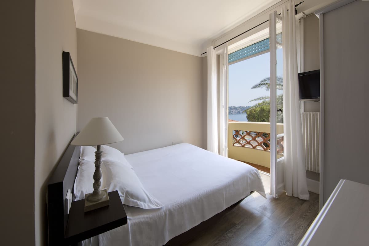 A hotel room with a bed, a bedside table with a lamp, a TV mounted on the wall, and a balcony with a view of the sea.