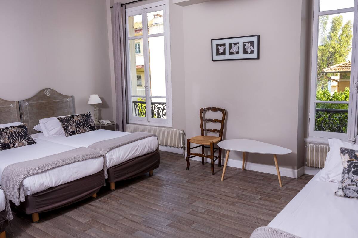 A bedroom with two beds, a chair, a table, and a window with a view of buildings and greenery.