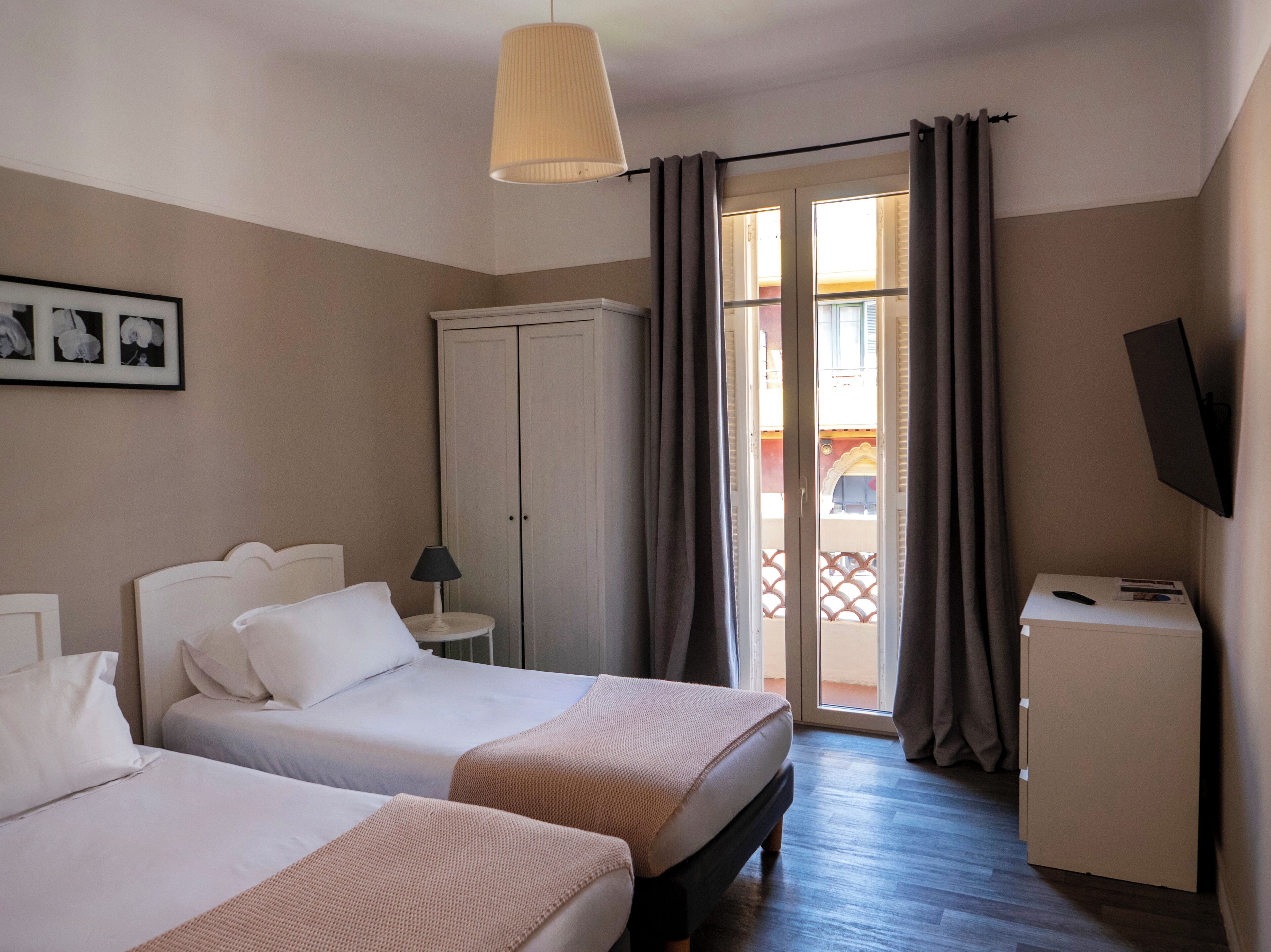 A hotel room with two beds, a wardrobe, a desk with a TV, and a balcony with curtains.