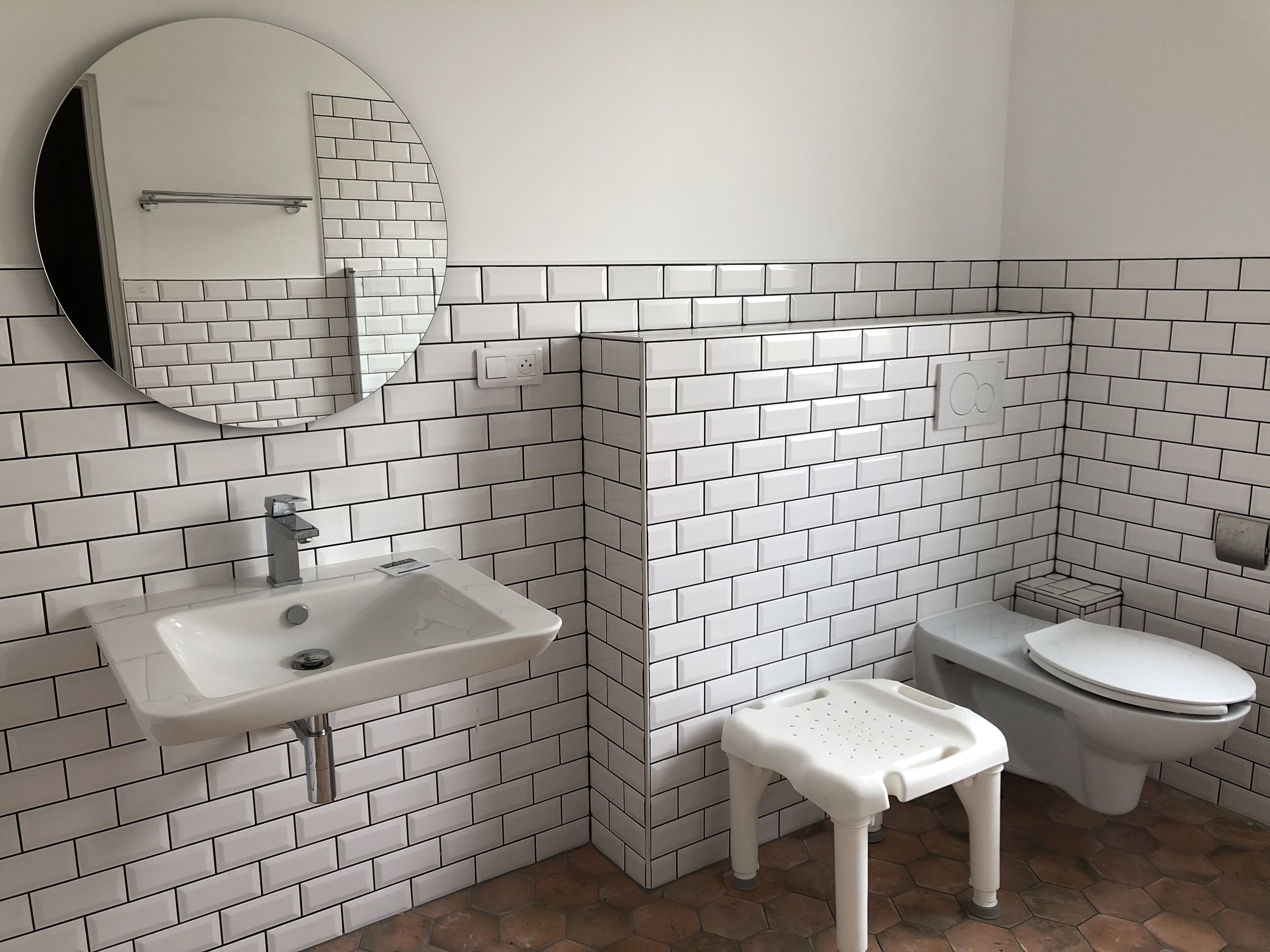 A bathroom with white subway tiles, a round mirror, a sink, a toilet, and a small white stool.