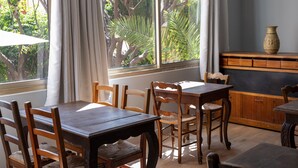 A dining area with a wooden table and chairs, a window with curtains, and a view of trees and greenery outside. - Hôtel Le Provençal (Villefranche-sur-Mer)