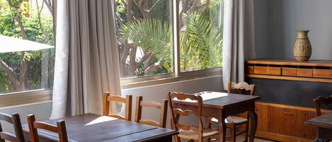 A dining area with a wooden table and chairs, a window with curtains, and a view of trees and greenery outside.