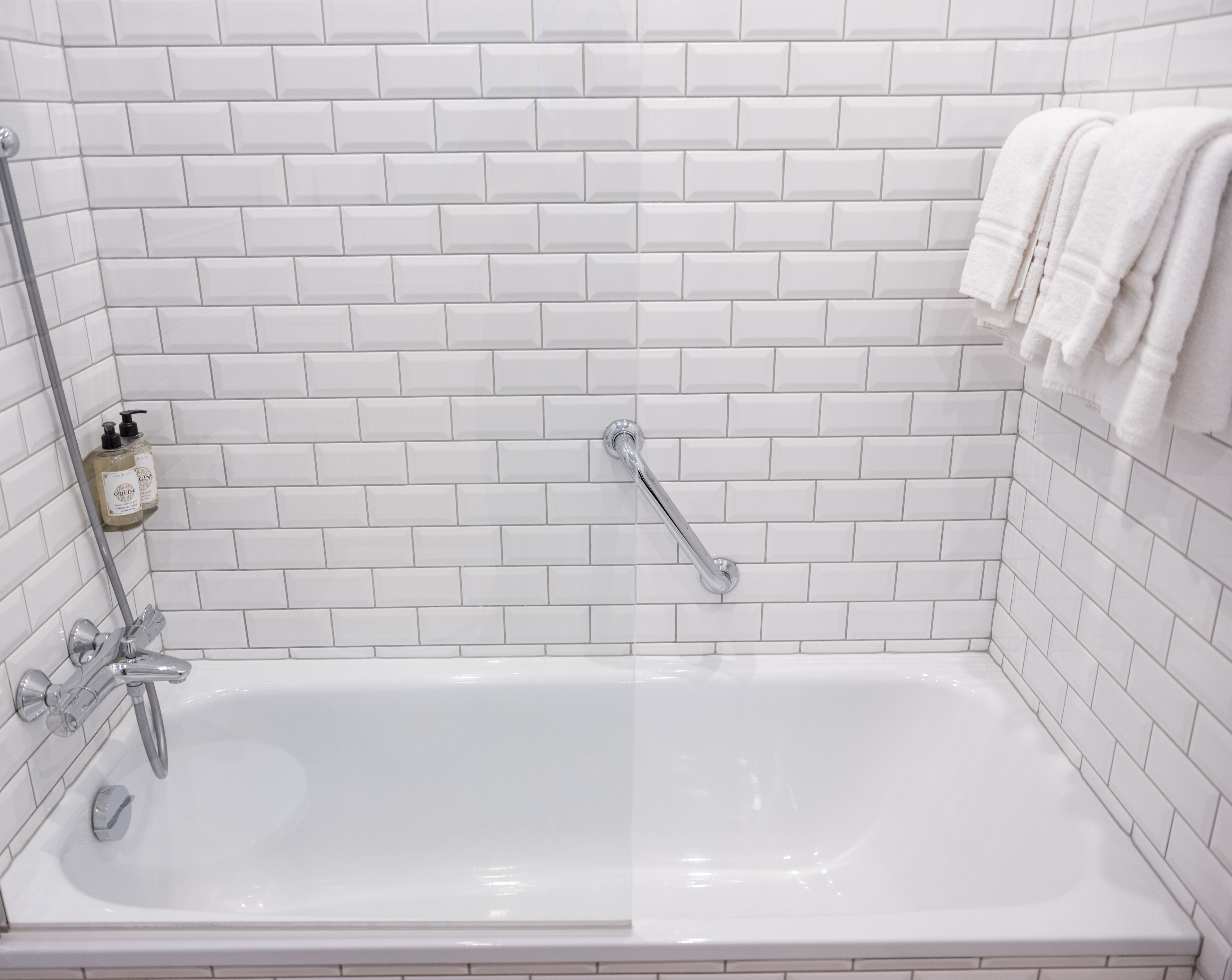 Une salle de bain aux murs recouverts de carreaux blancs, avec une baignoire, une douchette et une bouteille de shampoing biologique.