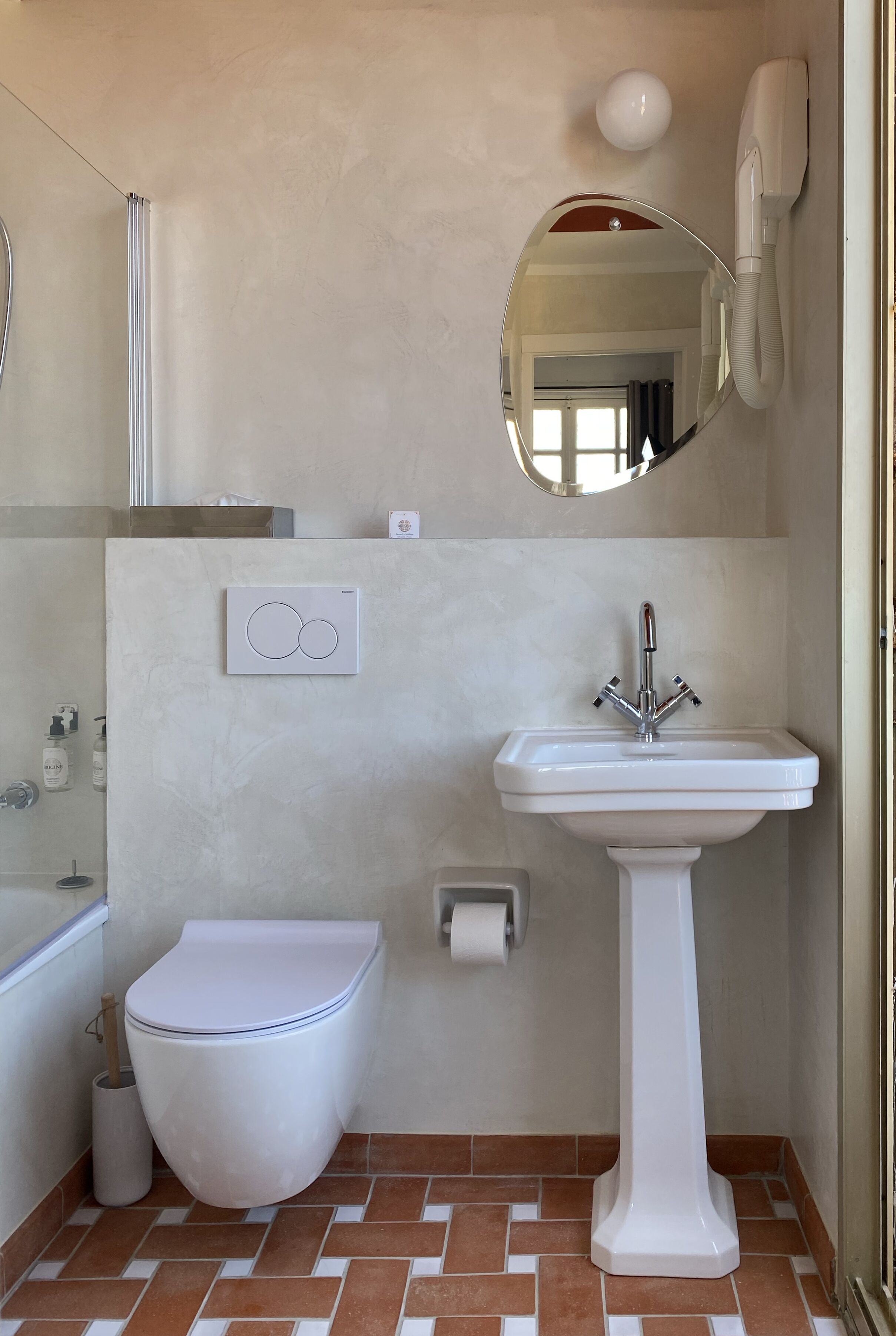 Un baño con inodoro blanco, lavamanos pedestal, espejo, cabezal de ducha manual y secador de pelo de pared.