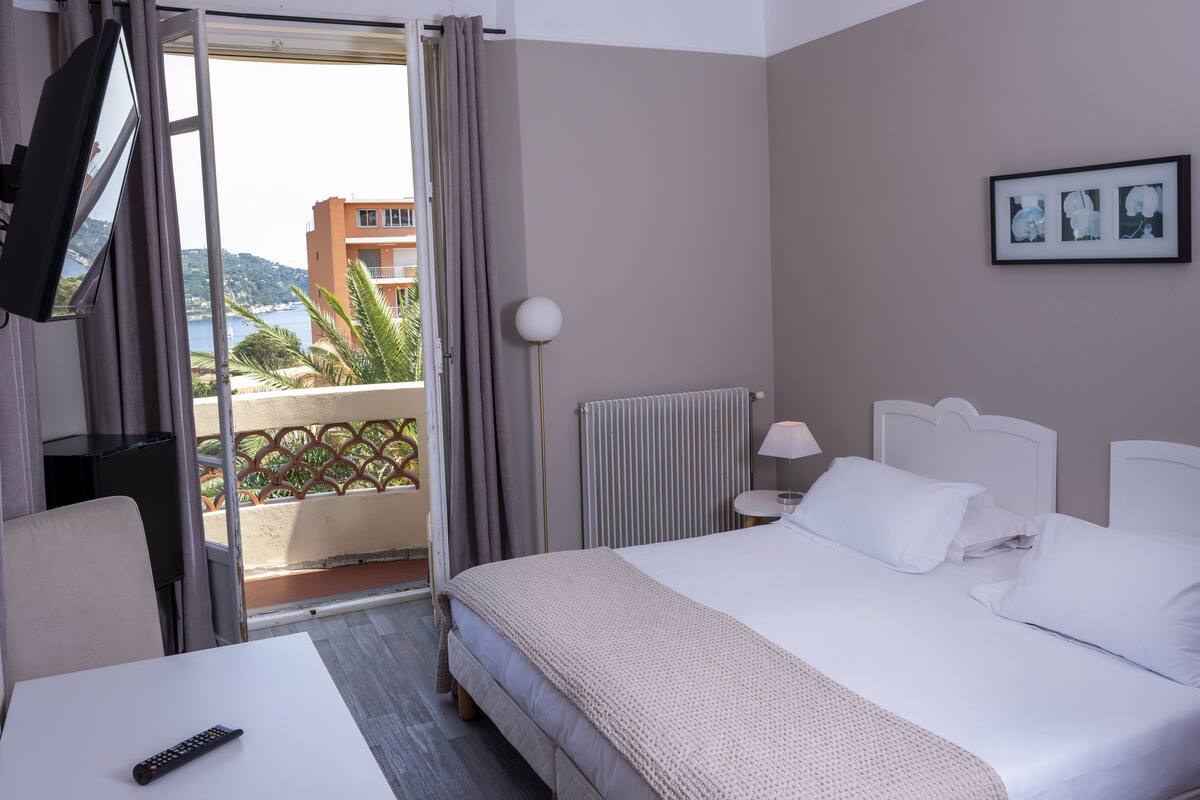 A hotel room with a bed, a TV, a desk, and a view of the sea.