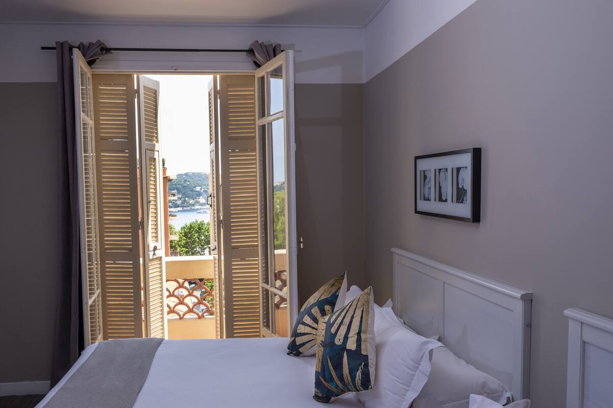 A hotel room with a bed, pillows, a framed picture on the wall, and an open balcony door.
