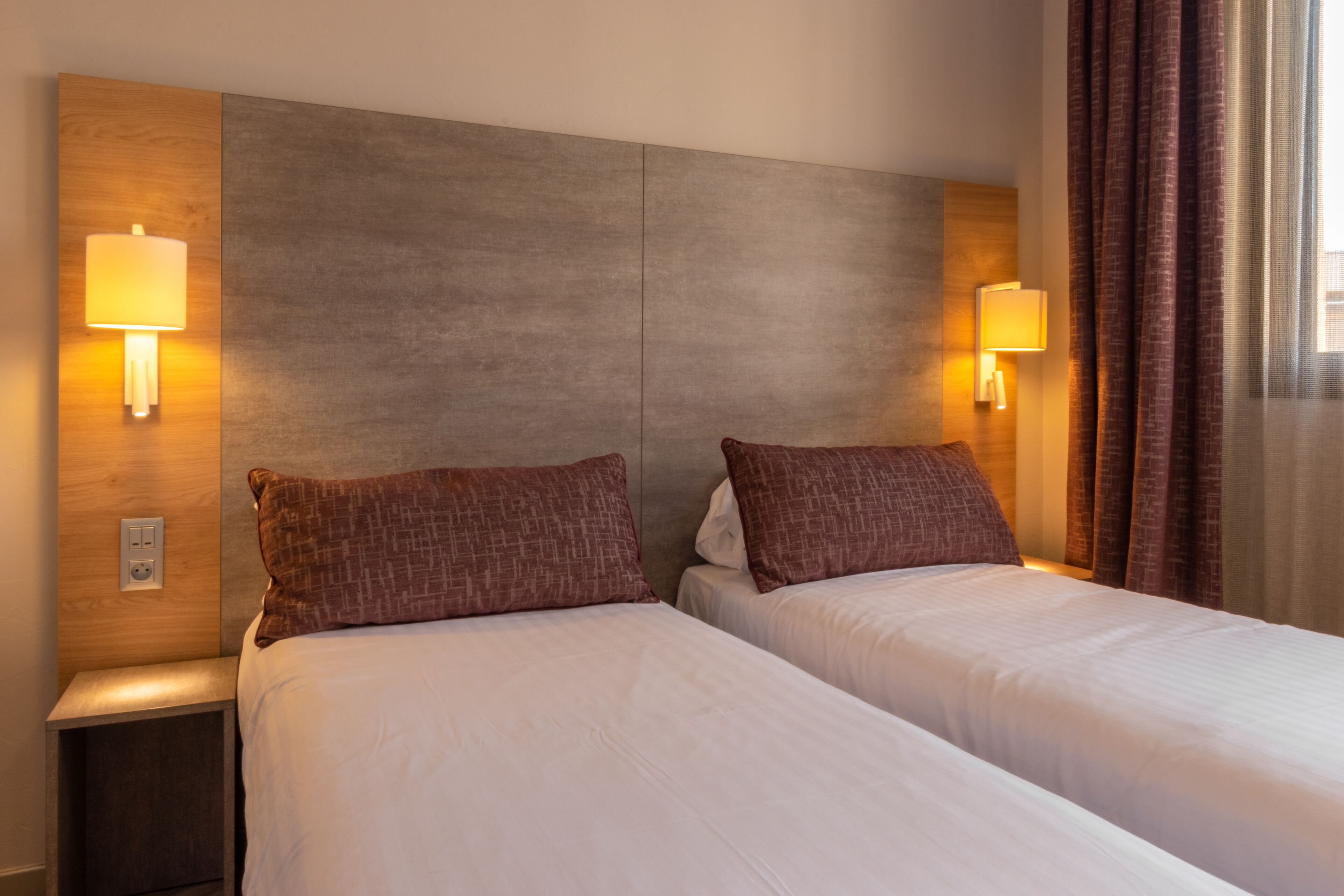 Habitación de hotel con dos camas, cabecero de madera, mesitas de noche, lámparas de pared y un ventanal con cortinas.