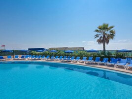 Una piscina con sillas reclinables azules y sombrillas, rodeada de vegetación y un cielo despejado.