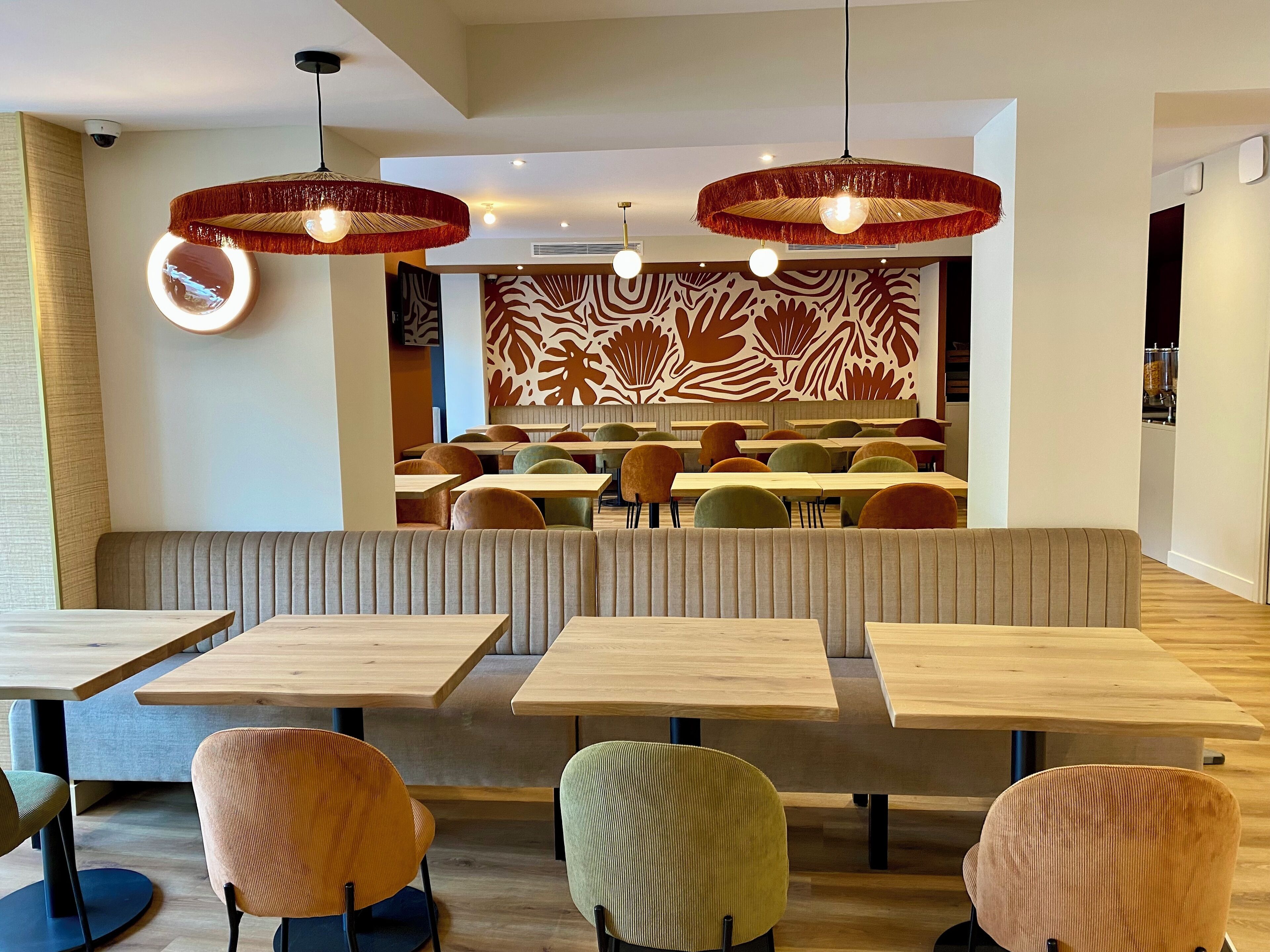 Interior de restaurante moderno con mesas de madera, sillas coloridas, pared con diseño y lámparas colgantes.