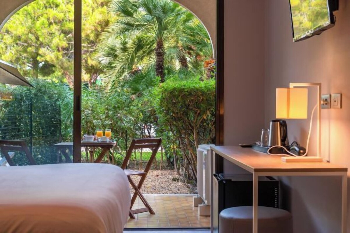 A room with a bed, a desk, a chair, and a view of a garden.
