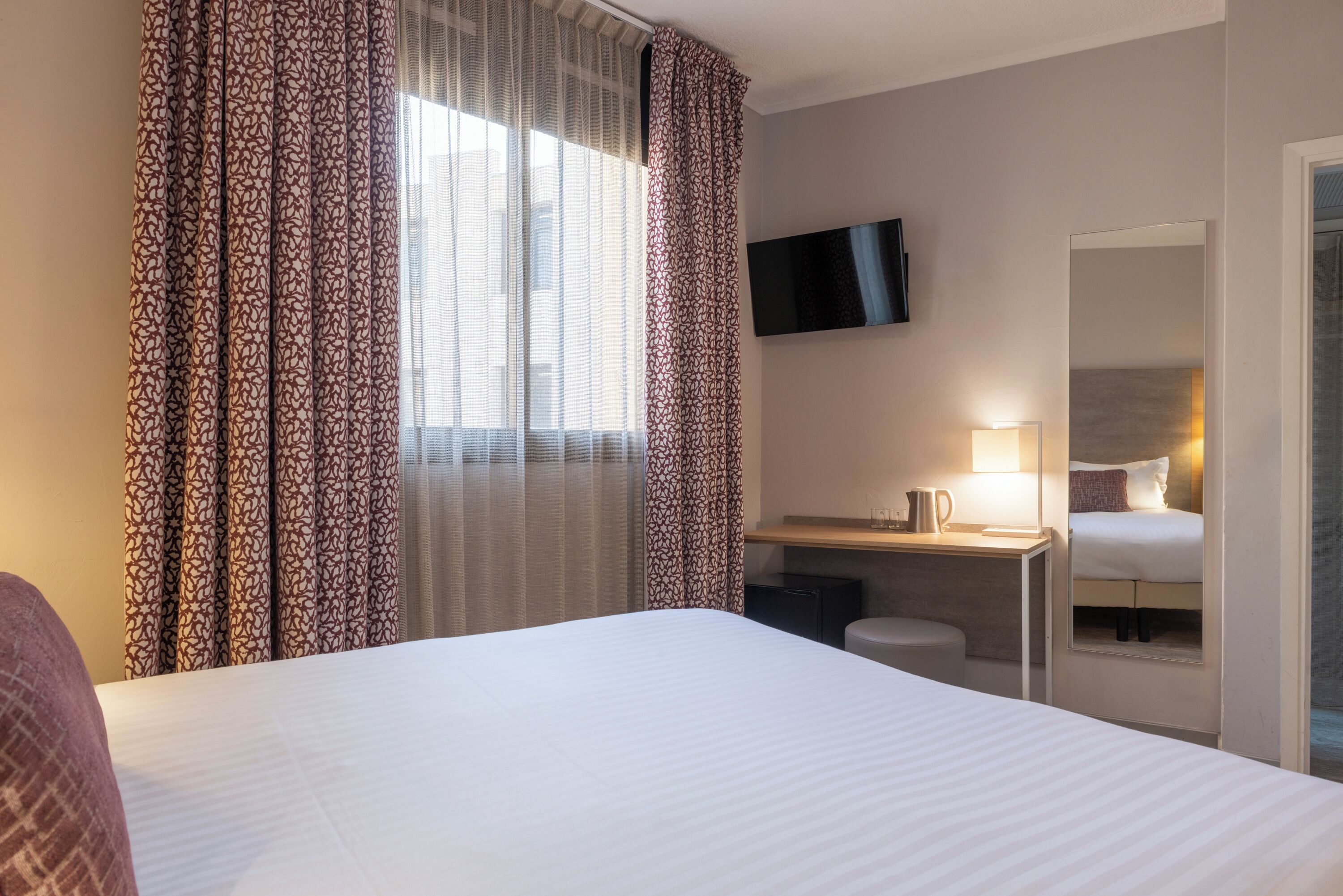 A hotel room with a large bed, a desk with a lamp, a TV mounted on the wall, and a window with patterned curtains.