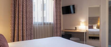 A hotel room with a large bed, a desk with a lamp, a TV mounted on the wall, and a window with patterned curtains.