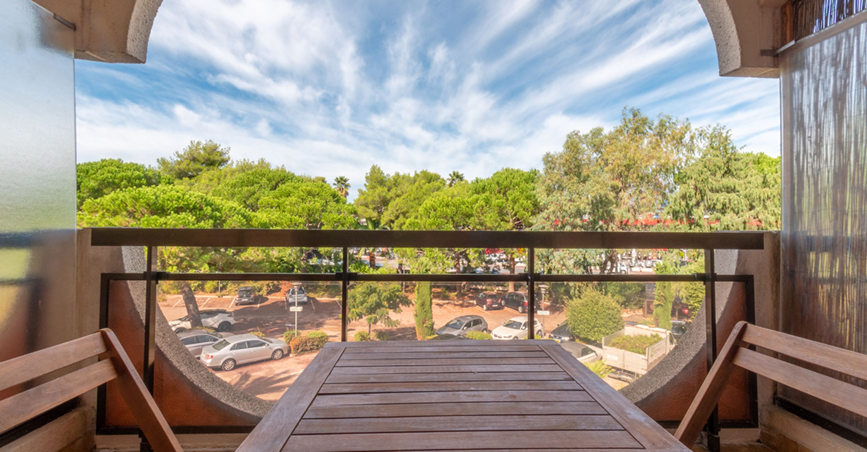 A balcony with a wooden table and benches, overlooking a parking area and trees.