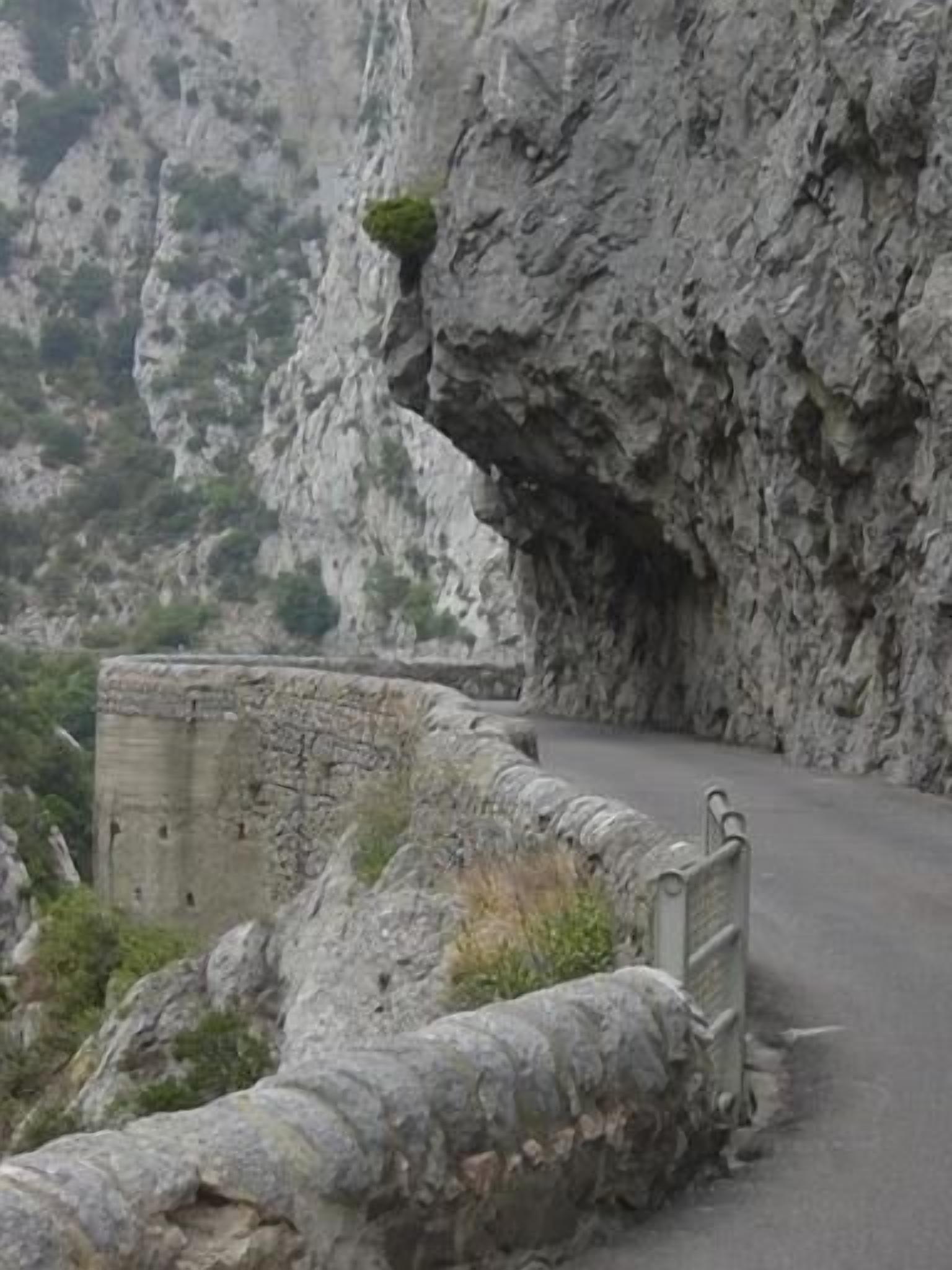 Una carretera de montaña estrecha y serpenteante, bordeada por muros de piedra y terreno rocoso.
