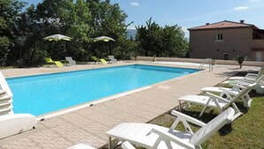Una piscina con sillas de descanso y sombrillas, rodeada de vegetación y un edificio.