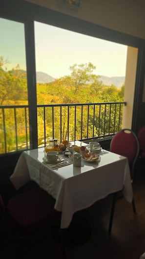 Une table à manger dressée avec des aliments pour le petit-déjeuner, et une vue sur un paysage montagneux à travers la rambarde d’un balcon.