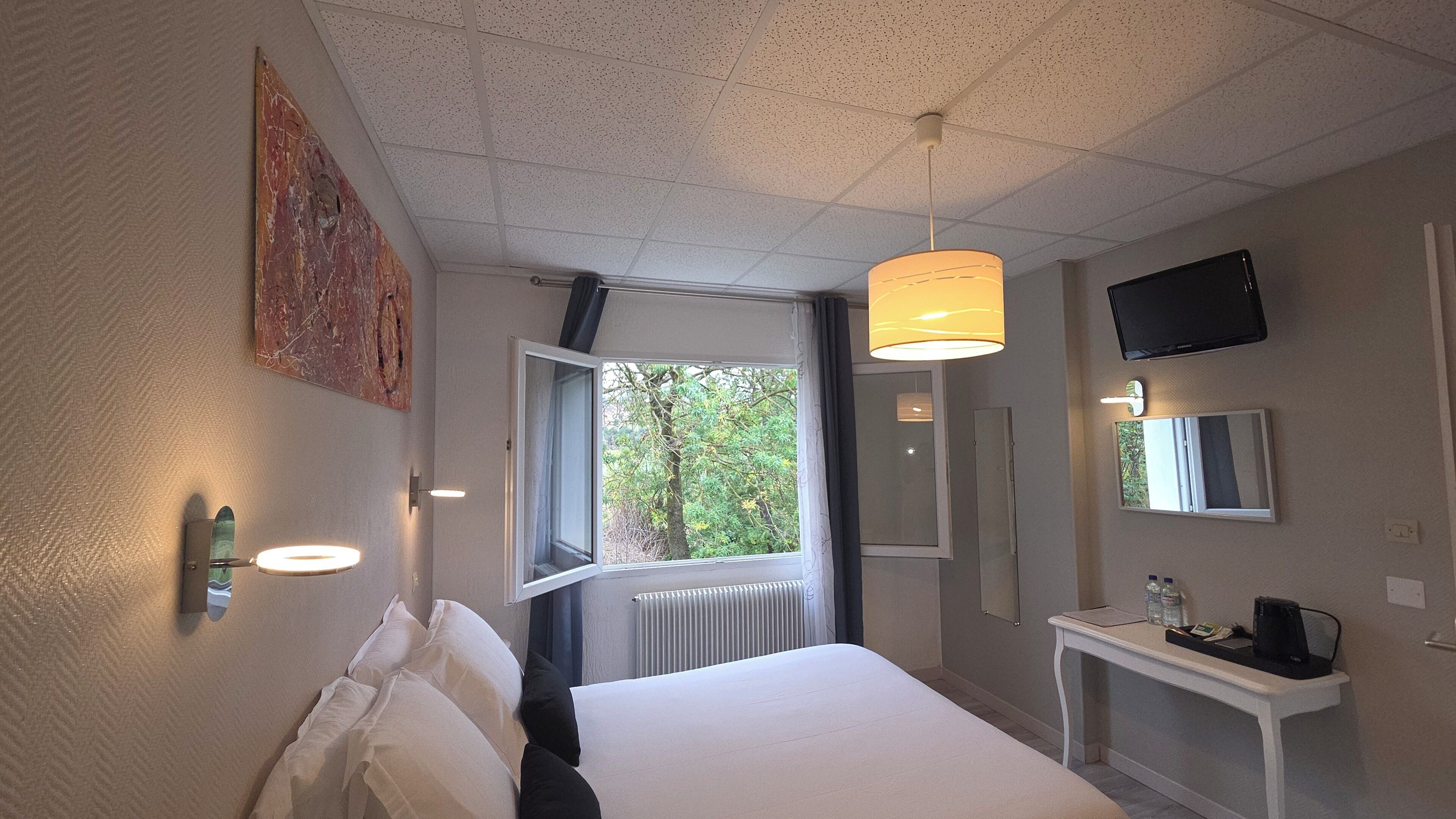 Une chambre d’hôtel équipée d’un lit, d’une table de chevet, d’une lampe, d’une fenêtre et d’un téléviseur fixé au mur.