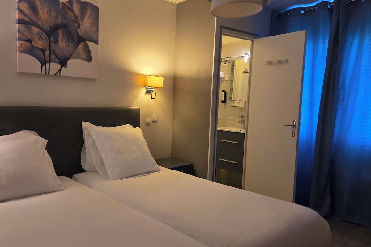 Habitación de hotel con cama doble, mesita de noche, lámpara de pared, puerta y baño visible a través de la puerta.