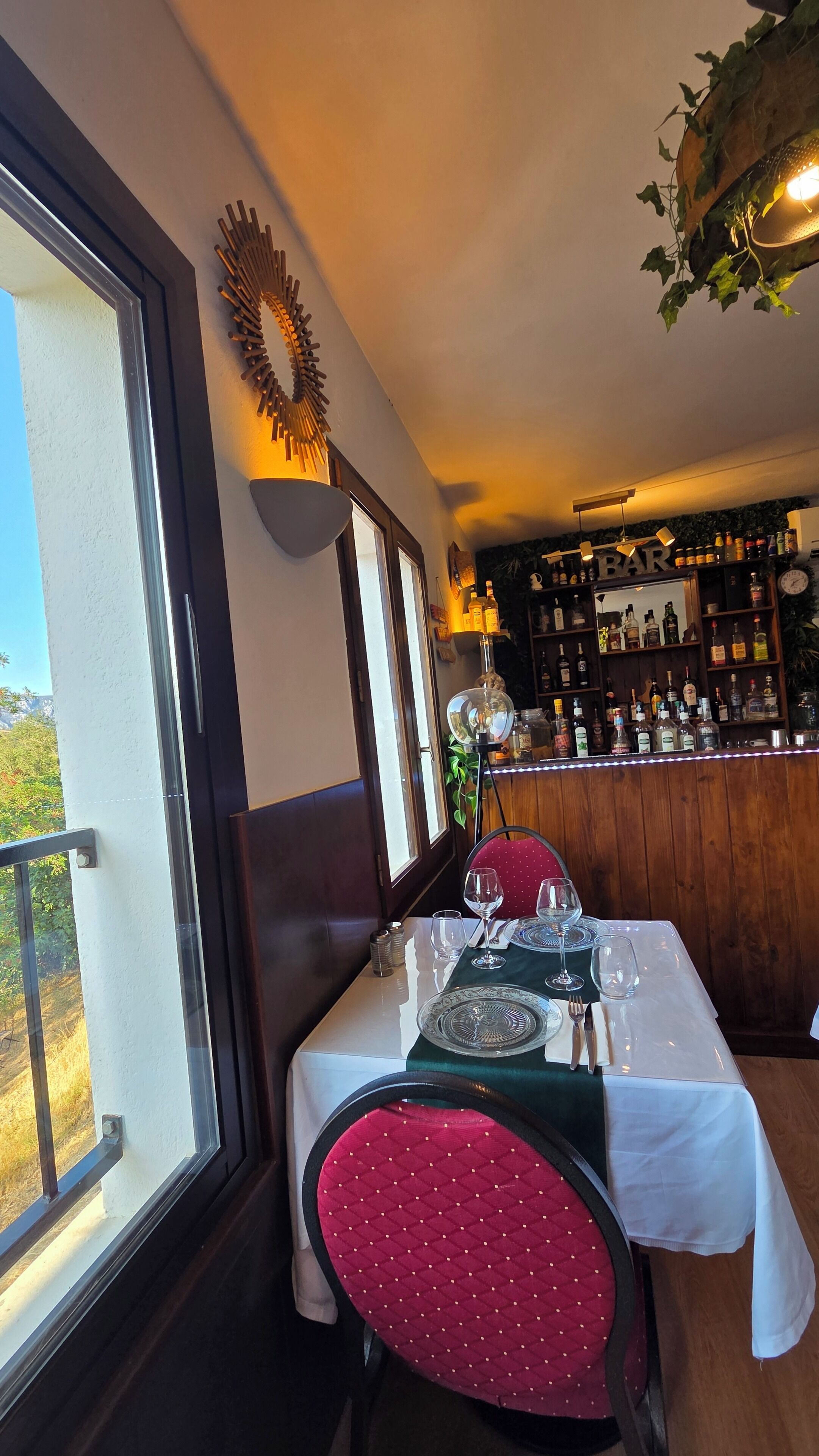 A dining area with a table set for two, a bar in the background, and a view through the window.