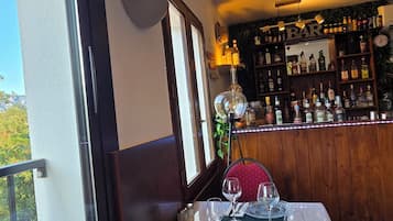 A dining area with a table set for two, a bar in the background, and a view through the window.
