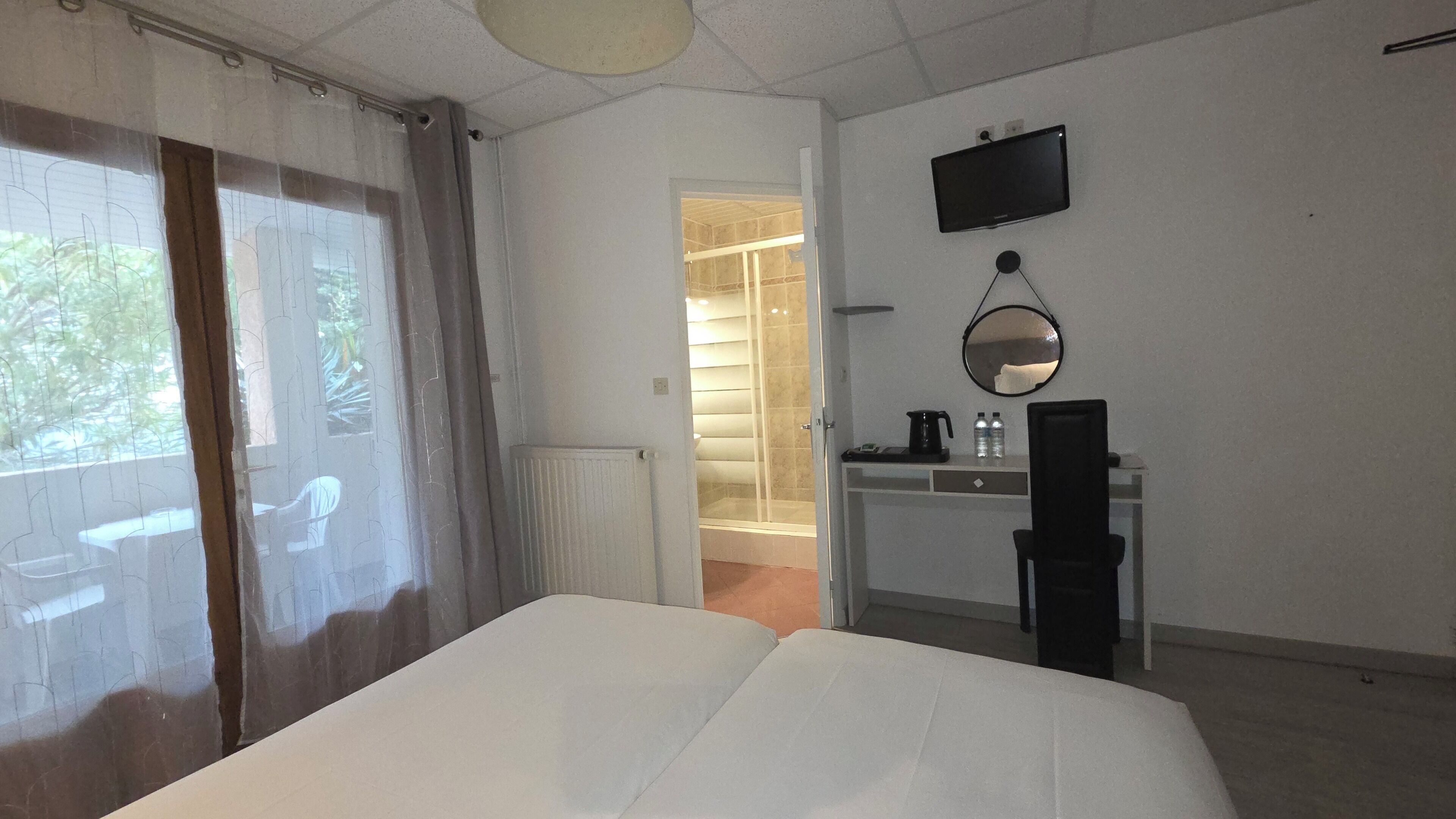 A hotel room with a bed, a desk with a computer, a mirror, and a shower area.