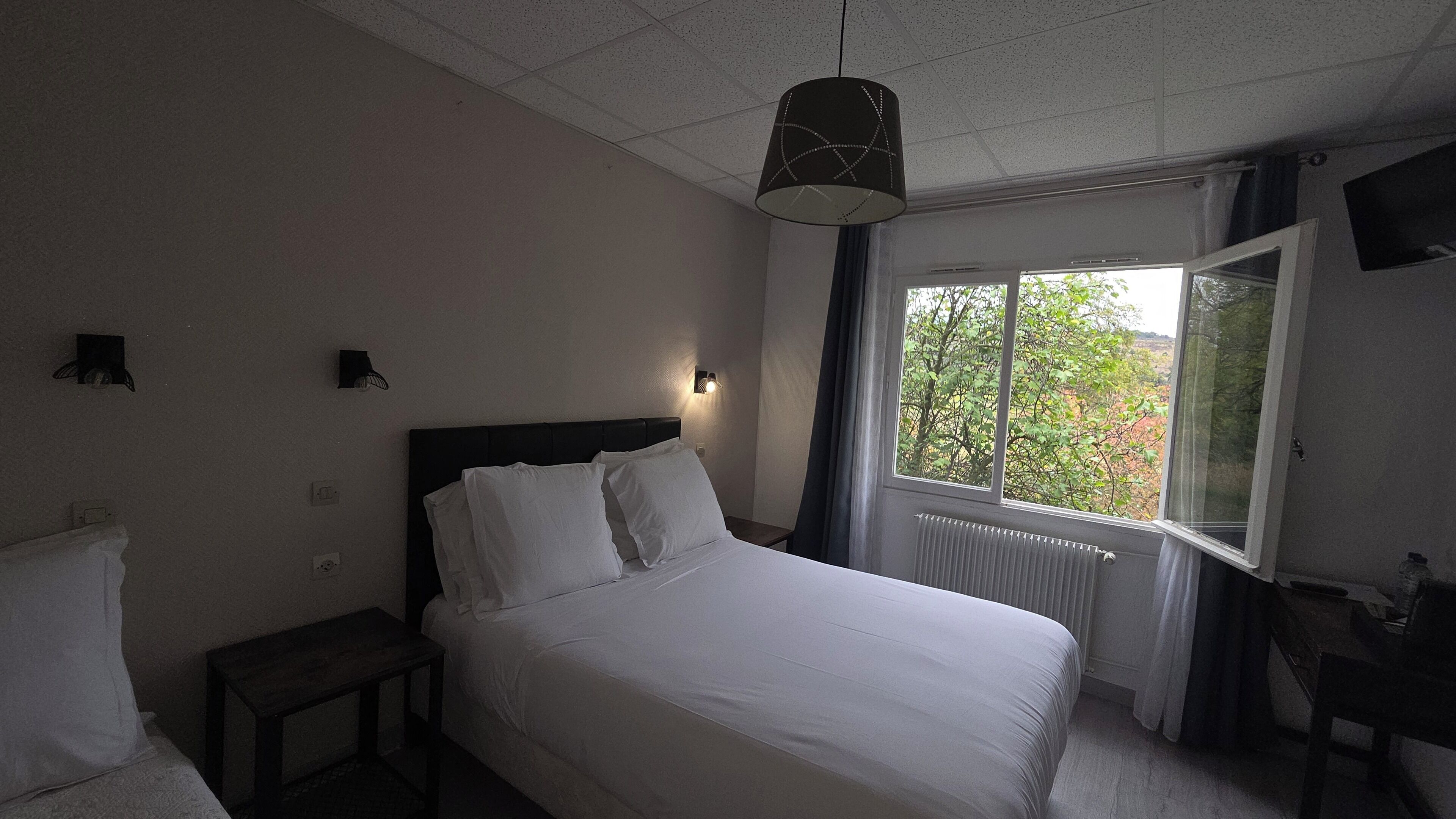 A hotel room with a bed, bedside table, lamp, window with curtains, and a view of trees.