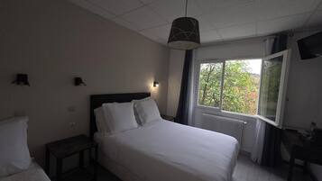 A hotel room with a bed, bedside table, lamp, window with curtains, and a view of trees.