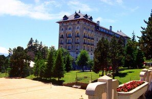 A large, multi-story hotel with a classic architectural style, surrounded by a well-maintained garden and trees.