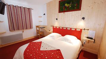 Ein Schlafzimmer mit einem Holz-Kopfteil, einer rot-weißen Bettdecke, einer kuscheligen Decke mit Herzmuster in Rot und Weiß, einem Nachttisch aus Holz mit einer Lampe, einem Holzstuhl und einem an der Wand befestigten Regal mit Büchern.