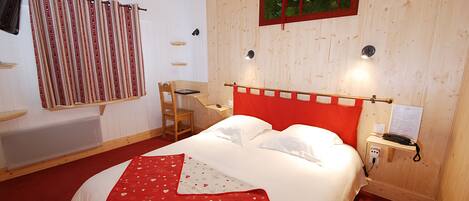 A bedroom with a wooden headboard, a red and white bedspread, a red and white heart-patterned throw blanket, a wooden nightstand with a lamp, a wooden chair, and a wall-mounted shelf with books.