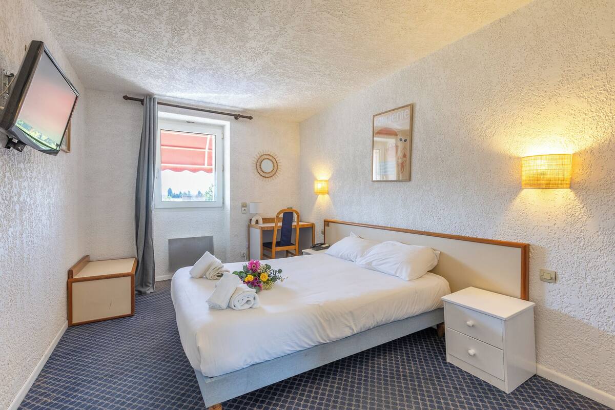 Hotellihuone, jossa on sänky, yöpöydät, työpöytä tuolilla, televisio ja ikkuna, jossa on verhot.