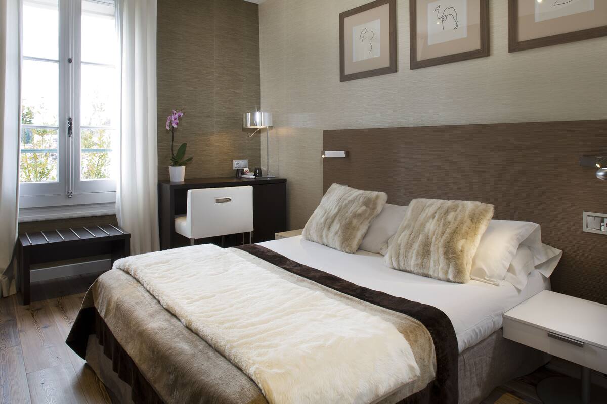 A bedroom with a large bed, a bedside table, a nightstand, a desk with a lamp, a potted plant, and framed artwork on the wall.