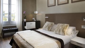 Une chambre avec un grand lit, une table de chevet, une commode, un bureau avec une lampe, une plante en pot et des œuvres d’art encadrées accrochées au mur.