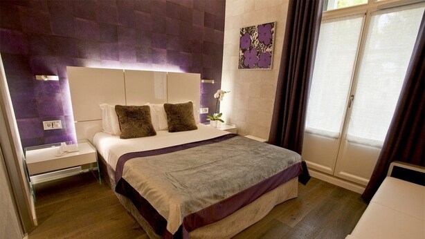 A modern bedroom with a large bed, a nightstand, a bedside table, and a window with curtains. - Hôtel des Arceaux (Montpellier)