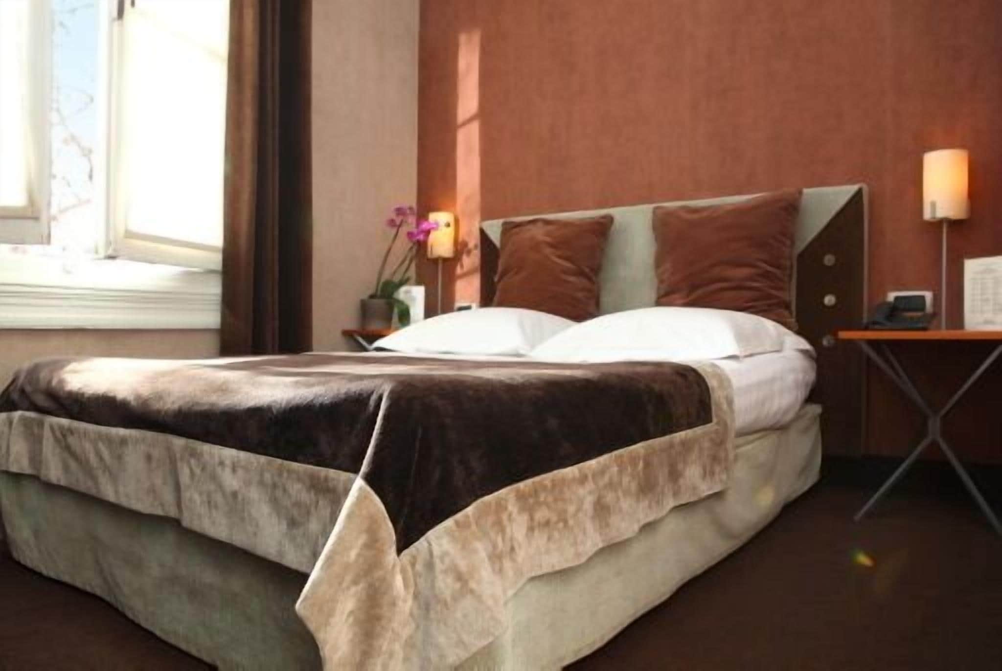 Een hotelkamer met een groot bed, een nachtkastje met een lamp en een raam met gordijnen.