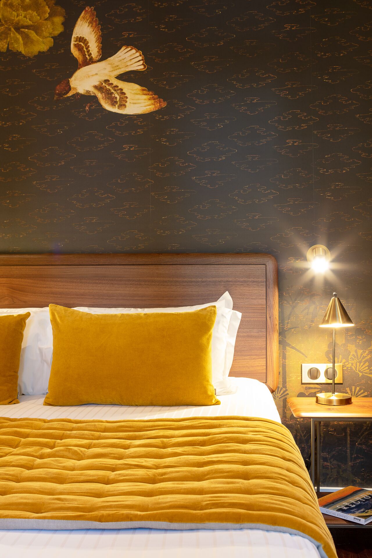 Una habitación de hotel con una cama, una mesita de noche, una lámpara y un mural de un pájaro en la pared.