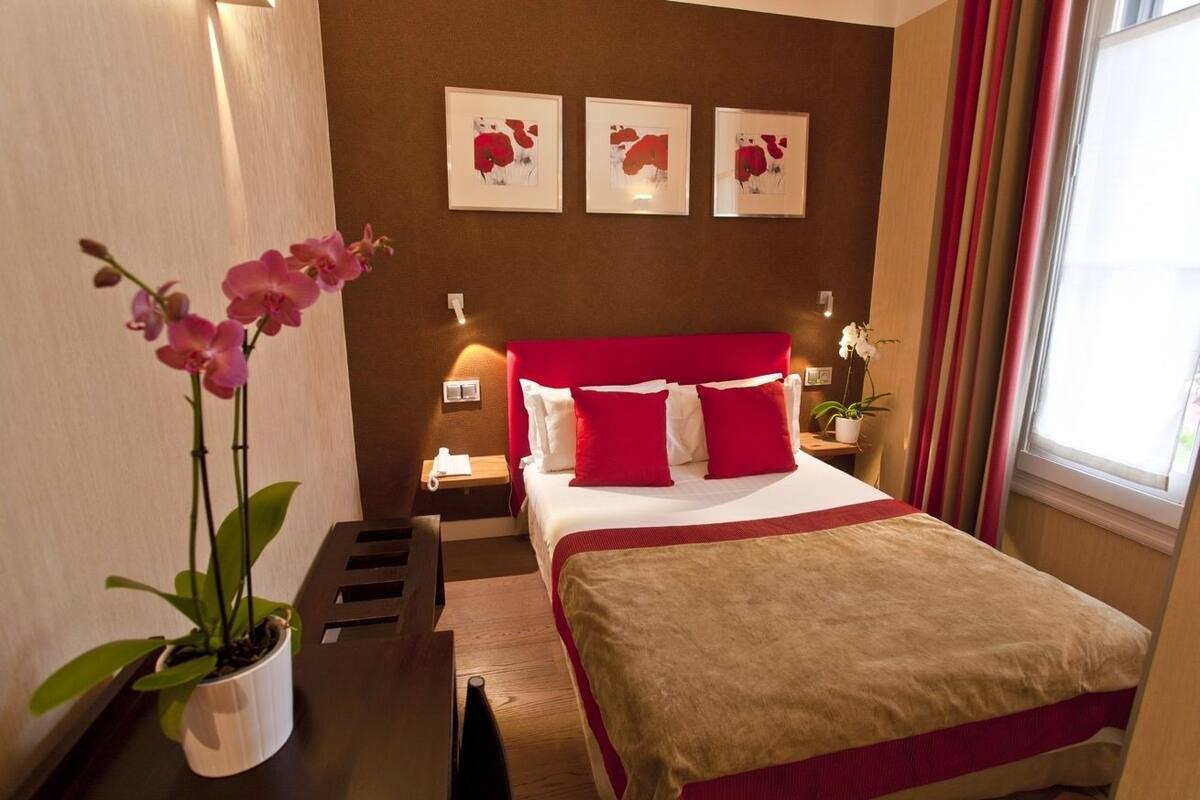 A hotel room with a large bed, red and white bedding, a nightstand with a lamp, and a potted orchid.