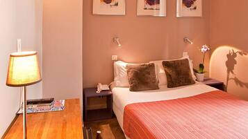 A hotel room with a bed, a bedside table with a lamp, a phone, and a small table with a magazine. There are three framed pictures on the wall above the bed.