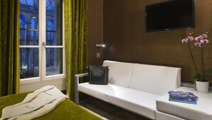 A hotel room with a white sofa, a small table with a plant and books, a window with curtains, and a flat-screen TV on the wall. - Hôtel des Arceaux (Montpellier)
